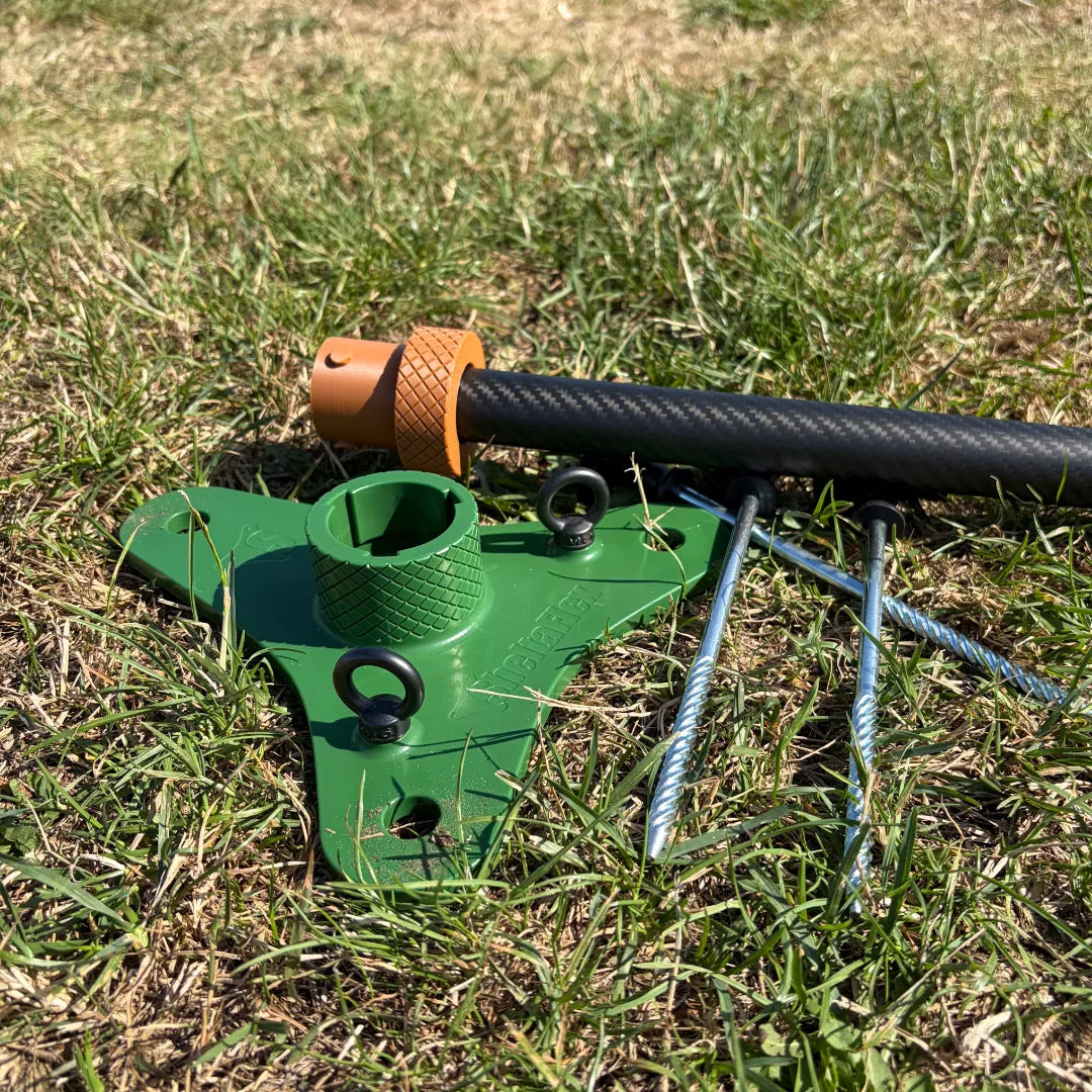 SheltaFlex FlexMount Base mit Erdanker auf einer Wiese, daneben liegt die passende Carbon-Teleskopstange.